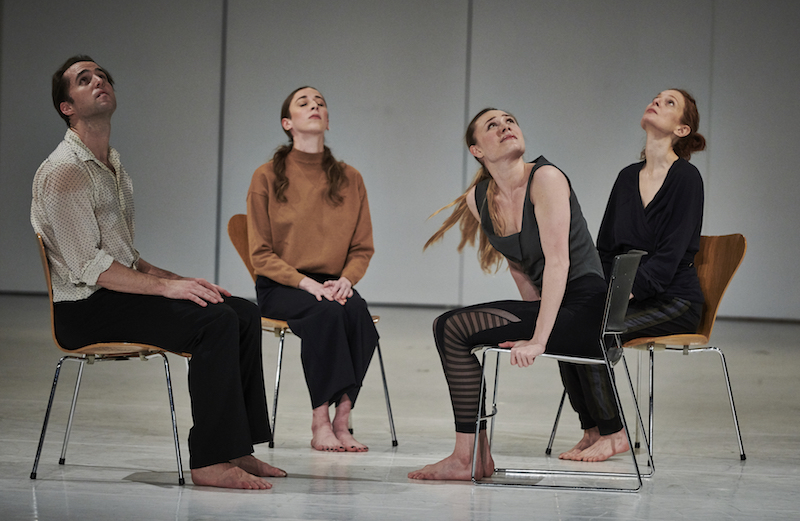 Four dancers sit in chairs with metal legs. Three look towards the ceiling and another tranquilly closes her eyes.