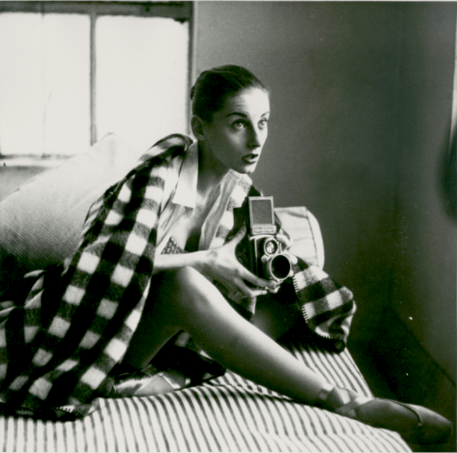 Tanaquil Le Clerq sitting on a bed with the covers pulled up around her and holding a polaroid camera.