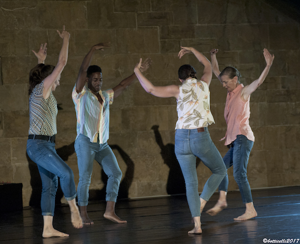 Molissa Fenley and three dancers stand in a circle with their arms stretched above their heads. She wears jeans and colorful patterned shirts.