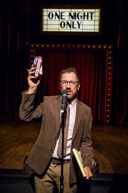 Robert wearing a suit talks into a microphone. He holds up a box of cracker jacks.