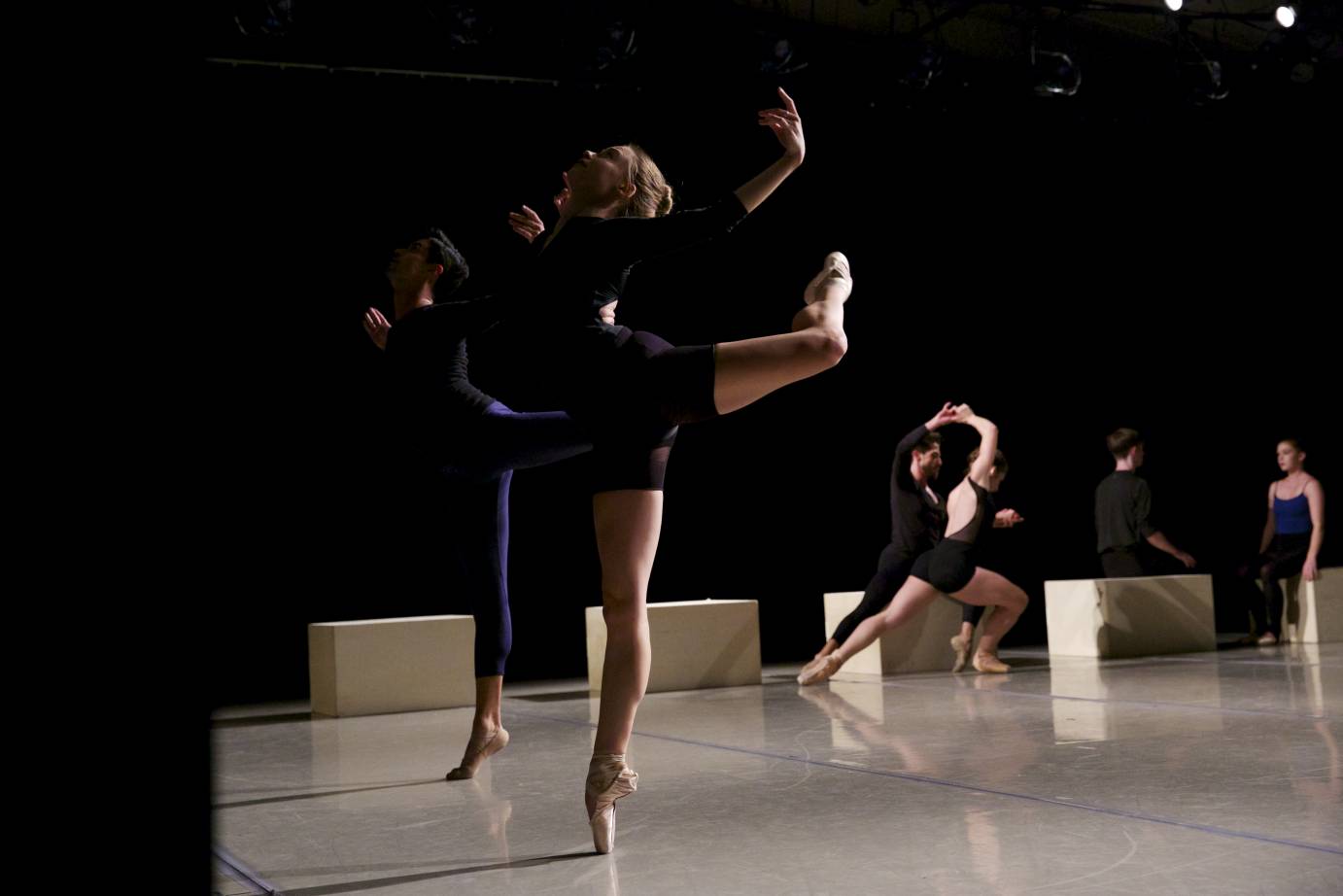Two people pose in attitude in relve on one side of the side while other dancers lunge and sit in the background.