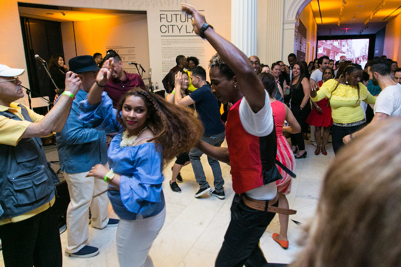 A crowd of people dance in the atrium of the Museum of the City of New York. A woman in blue blouse and white jeans is in the foreground. Her hair fans out as a man in a red vest spins her.