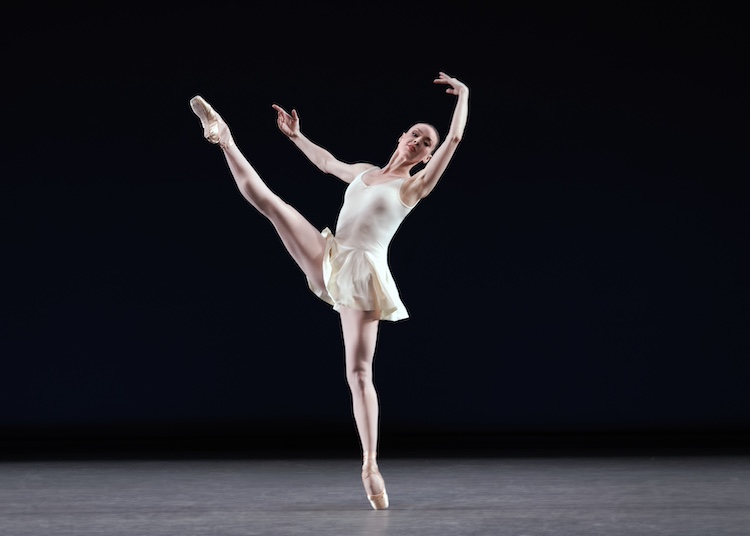 dancer on pointe, wearing pink tights and a white leotard with a minny skirt, kicks her leg hight forward
