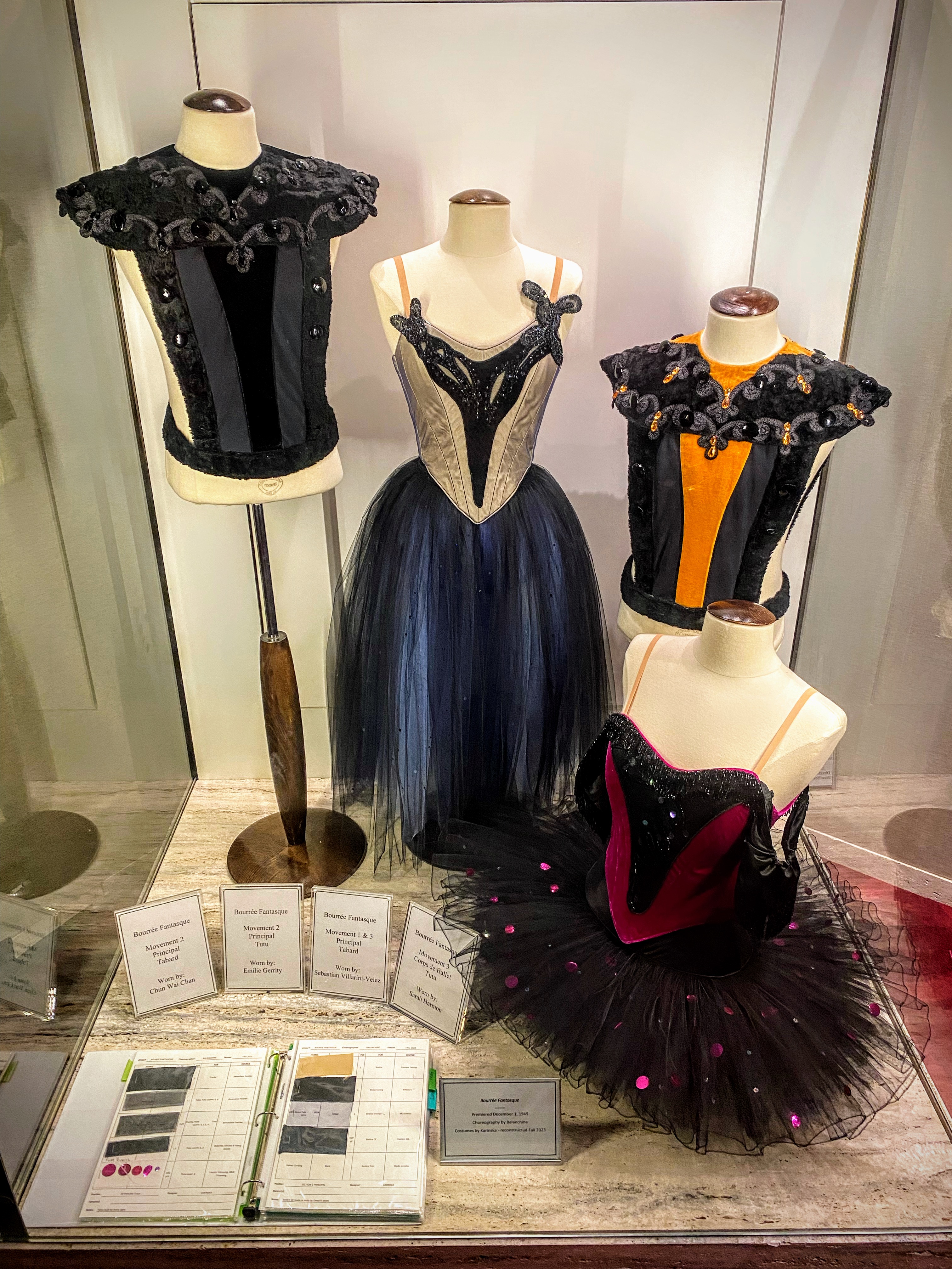 ballet bodices and tutus with a book of color swatches on display in the lobby of the Koch Theater. The colors for mens bodices are black and black and orange with intricate lace designs at the top. The tutus one romantic with a long skirt and one more classic with a short full skirt are in shades of cream and blue, and red and black respectively