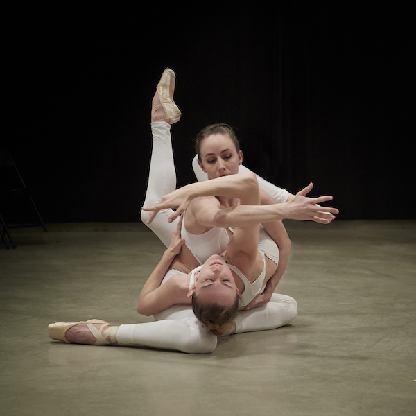 Two female dancers in white are interwined like pretzels on the floor. They both wear pointe shoes.