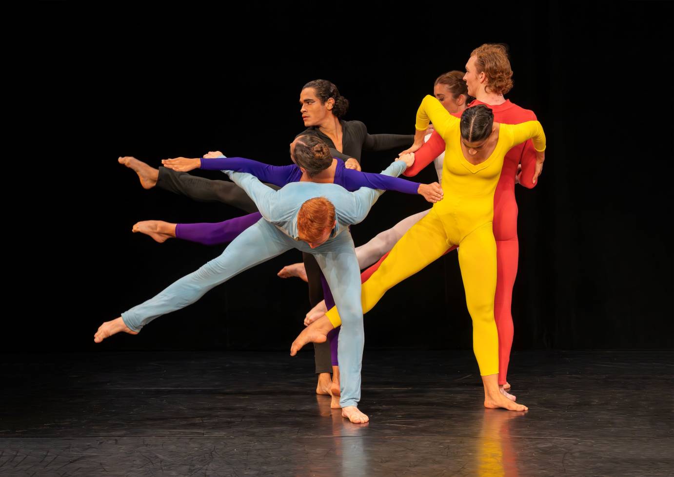 the company in striking  colorful unitards of purple, black, yellow, light blue, and orange cling together to form and off-center and highly energized human sculpture