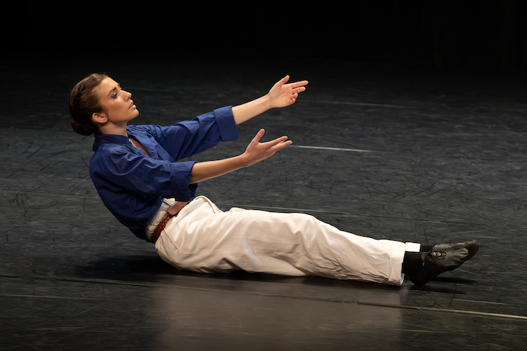 a woman in white pants and a blue shirt wearing black shoes sits on the floor reaching her arms out in yearning