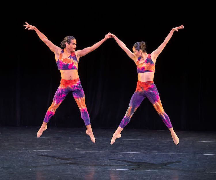 two dancers in bright kaleidoscopic tights and midriff bearing tops jump in the air holding hands... The duo creates a double x shape