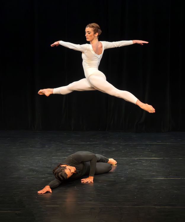 as a dancer in a black unitard lies on his side on the floor, a woman in a bright white unitard soars above him in a split jump also called a jete in the ballet world.