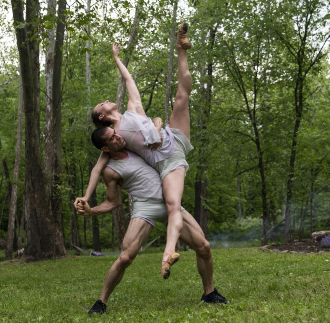 Against the backdrop of a woods in full greenery, a man lifts a woman who extends her leg highly to the side