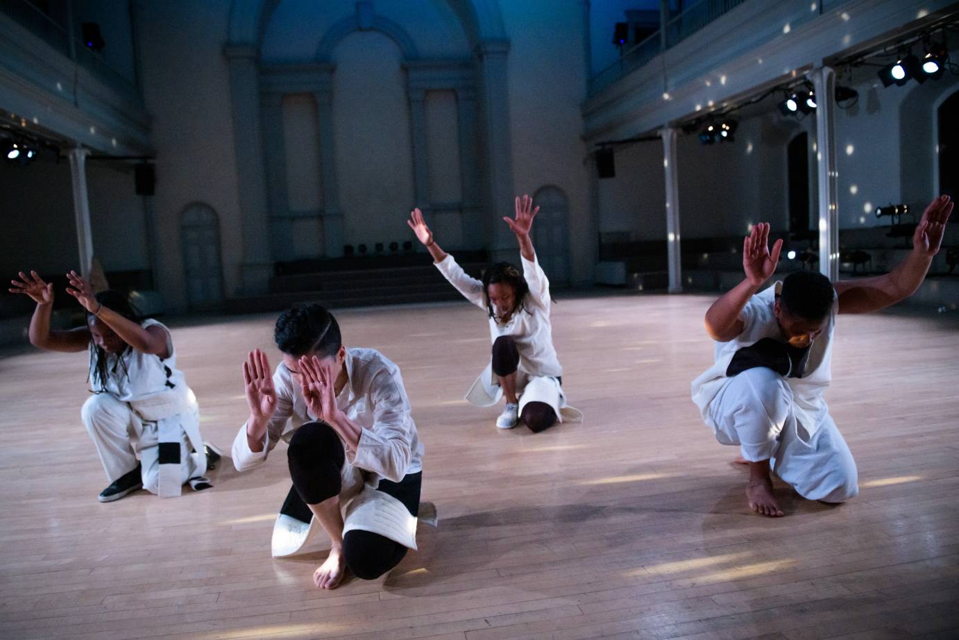 Four dancers in white kneel with their arms extended in front of them