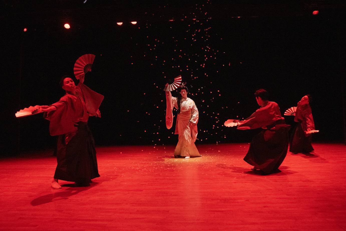 All performers are fluttering fans while standing on a red-bathed stage.