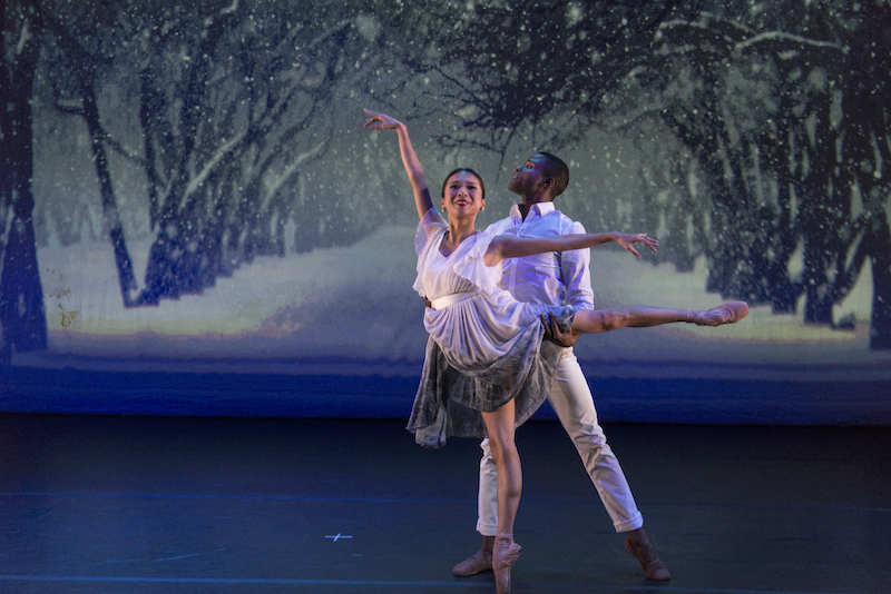 Two dancers pose majestically in white in front of a snowy tree-filled painted backdrop