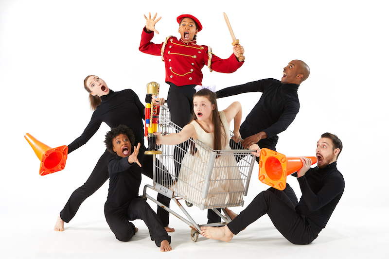 Dancers open their mouths wide as they pretend to tip a shopping cart. Some dancers are holding traffic cones. 
