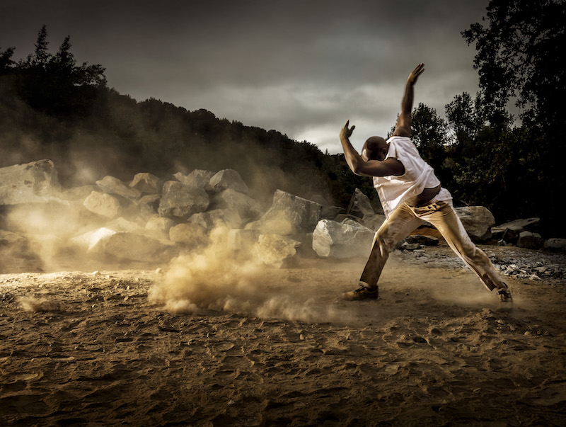 A dancer in all white strikes a pose on the dusty site of Andy Goldsworthy's stone tower project. His arm shields his face as dust swirls around him.