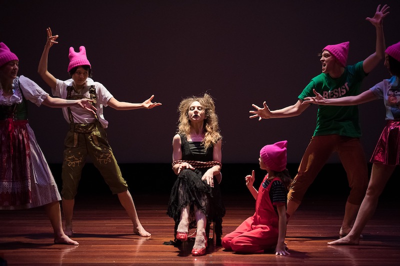 A group of performers pose around the actor who plays the witch. She wears a long black dress and sits in a chair. The others wear colorful casual clothes and Pink Pussie hats.