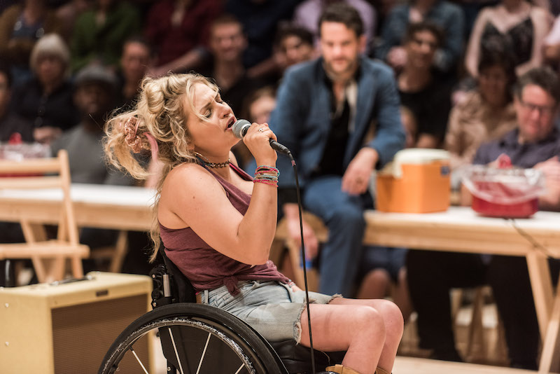 A woman with blonde hair closes her eyes singing into a microphone. She's seated in a wheelchair.