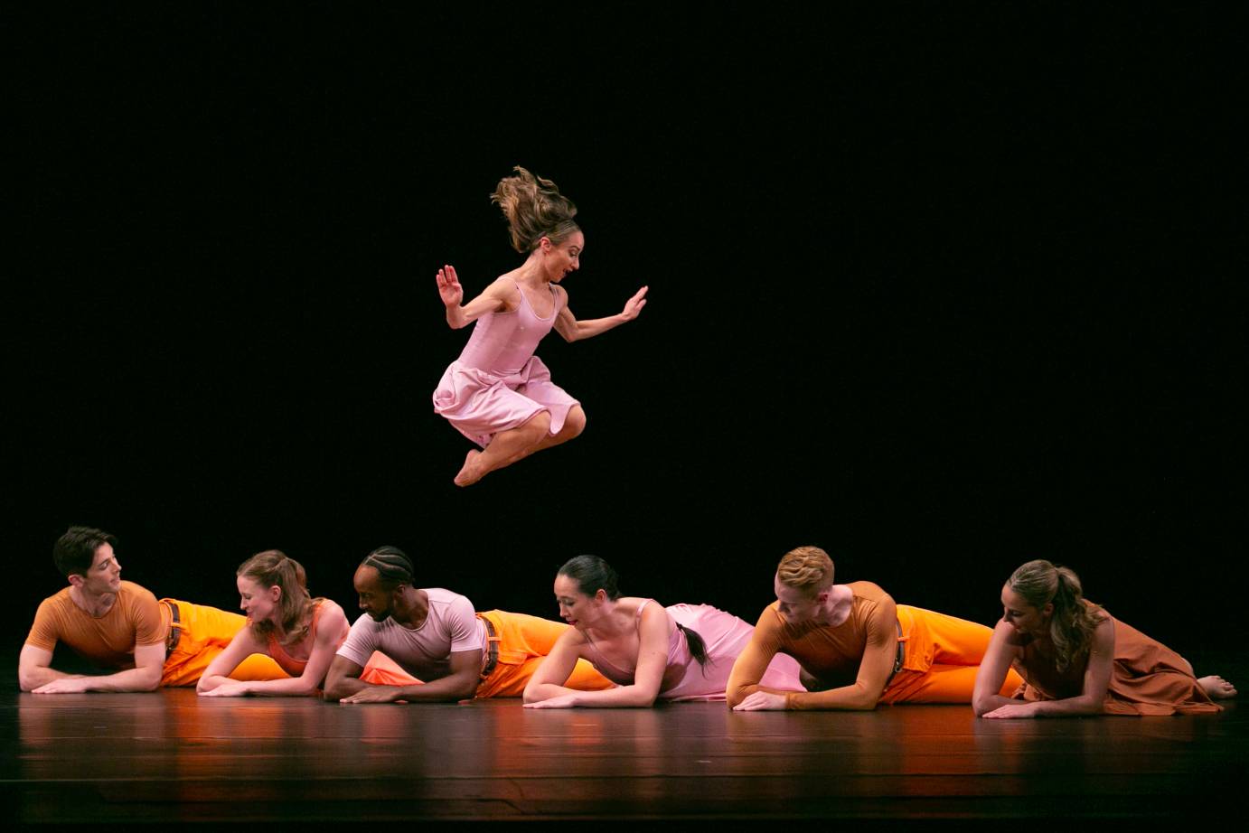 a woman in a pink dress joyously springs above a group of 6 dancers -men in orange and women in pink- lying on the floor. 