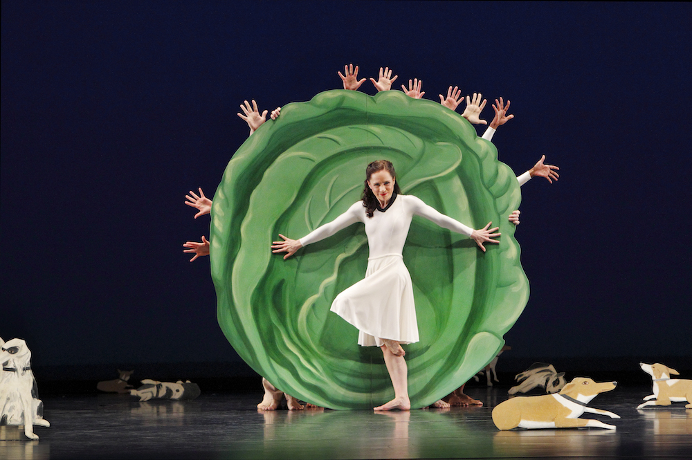 a light skinned female dancer wearing a lycra white dance dress holds a pose in front of a giant green cabbage cut out..surrounding her are cut outs of many kinds ofdogs