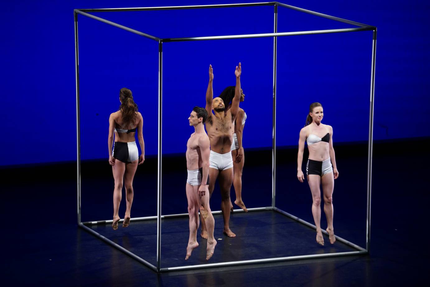dancers in black and white shorts and black and white bikinis jump upright in a minimalist version of a cube