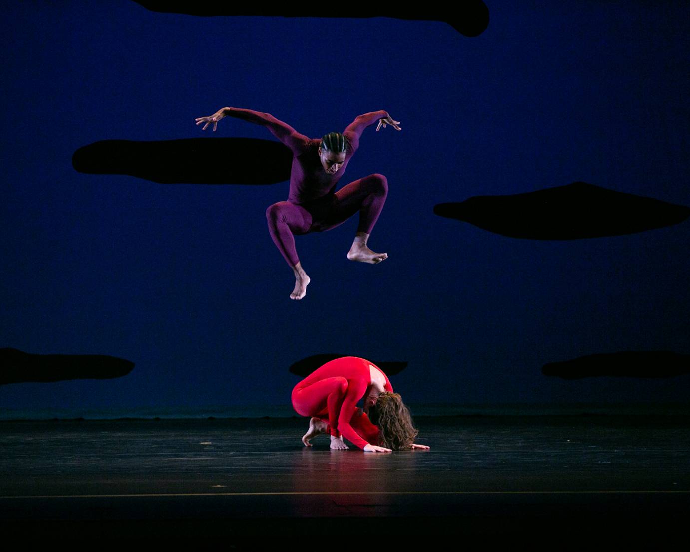 a Black man in a purple unitard jumps high above a woman in a red unitard crouched in a a ball like positon