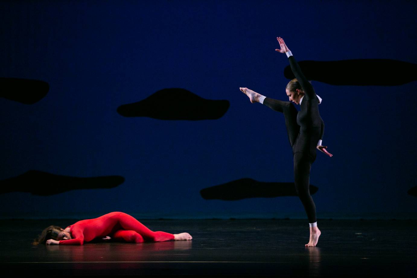 a woman in a red unitard lies on the floor as another woman dressed in a black unitard with a white collar and white cuffs hovers over her menacingly 