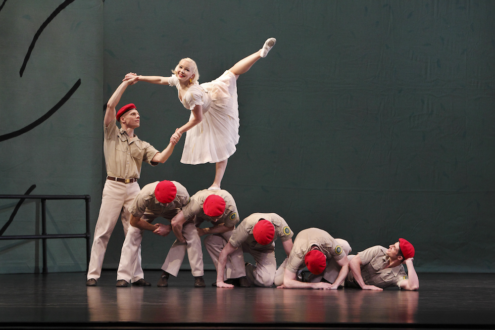 Men in uniforms with red berets form a cascading line from right to left- the man to the left is upright the rest of the line crouches... lower than lower.a young girl in a white sheer dress balances on the back of one of the men as the standing man holds her arms