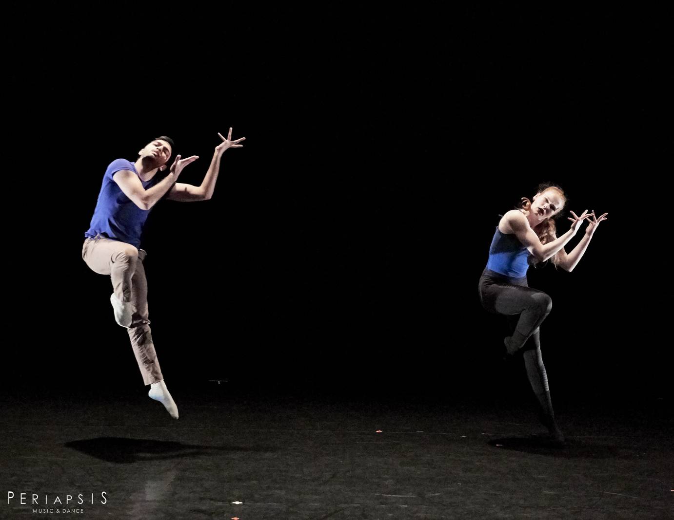 A man and a woman in blue tops jump with one leg touching their knee and splayed fingers