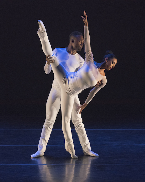 Two dancers in white. One dancer extends her leg high in the air as her torso drips to the side. Her partner stands behind her and helps her balance.