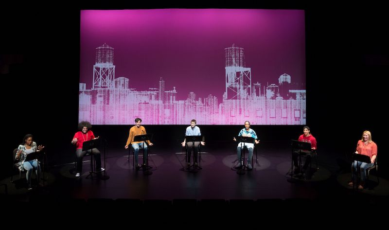 Students sit in chairs reading from the Undesirable element scripts in front of them that are placed on music stands. A magenta colored projection of a city skyline is displayed behind them.