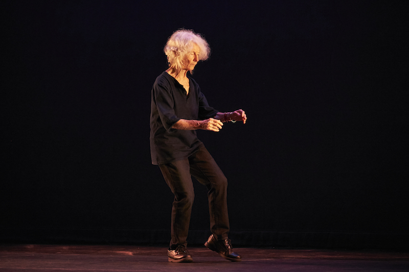 Simone Forti wears black slacks and shirt as she stands on one leg. Her two arms are in front of her.