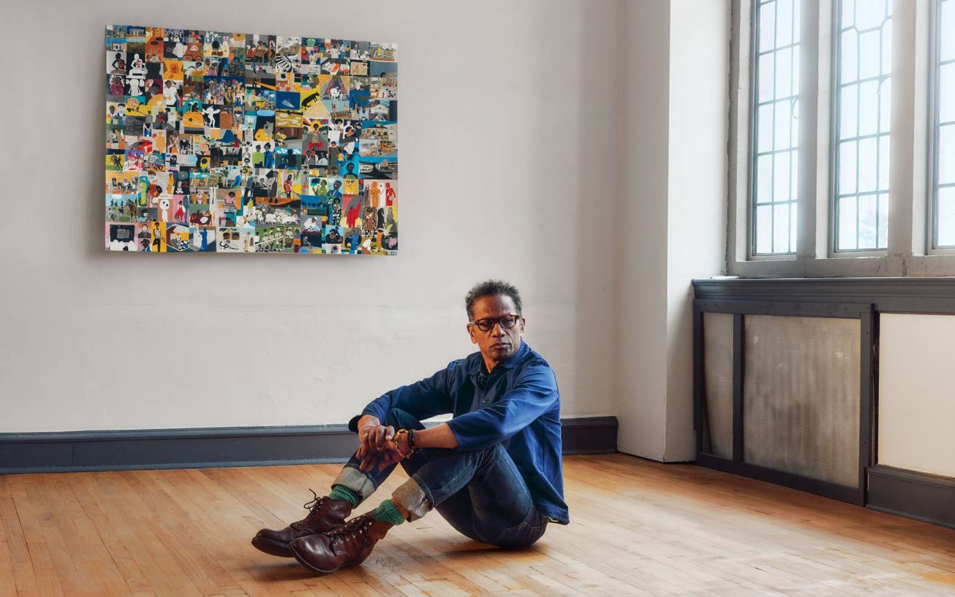 Ralph Lemon a Black Man dressed in denim wearing brown boots and colorful socks, sits on a wood floor of a studio space with his colorful mosaic painting in the background