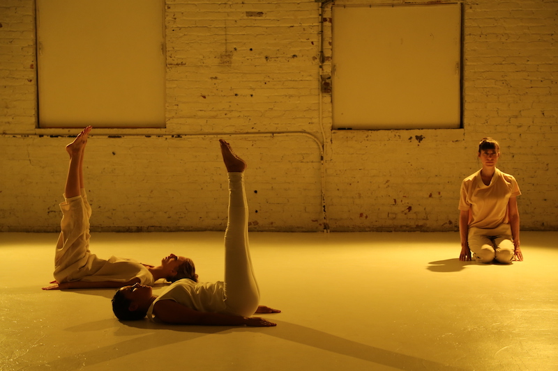 Two women lay on their backs with their legs raised to the ceiling. A third woman sits on her knees under a yellow soft glow. 