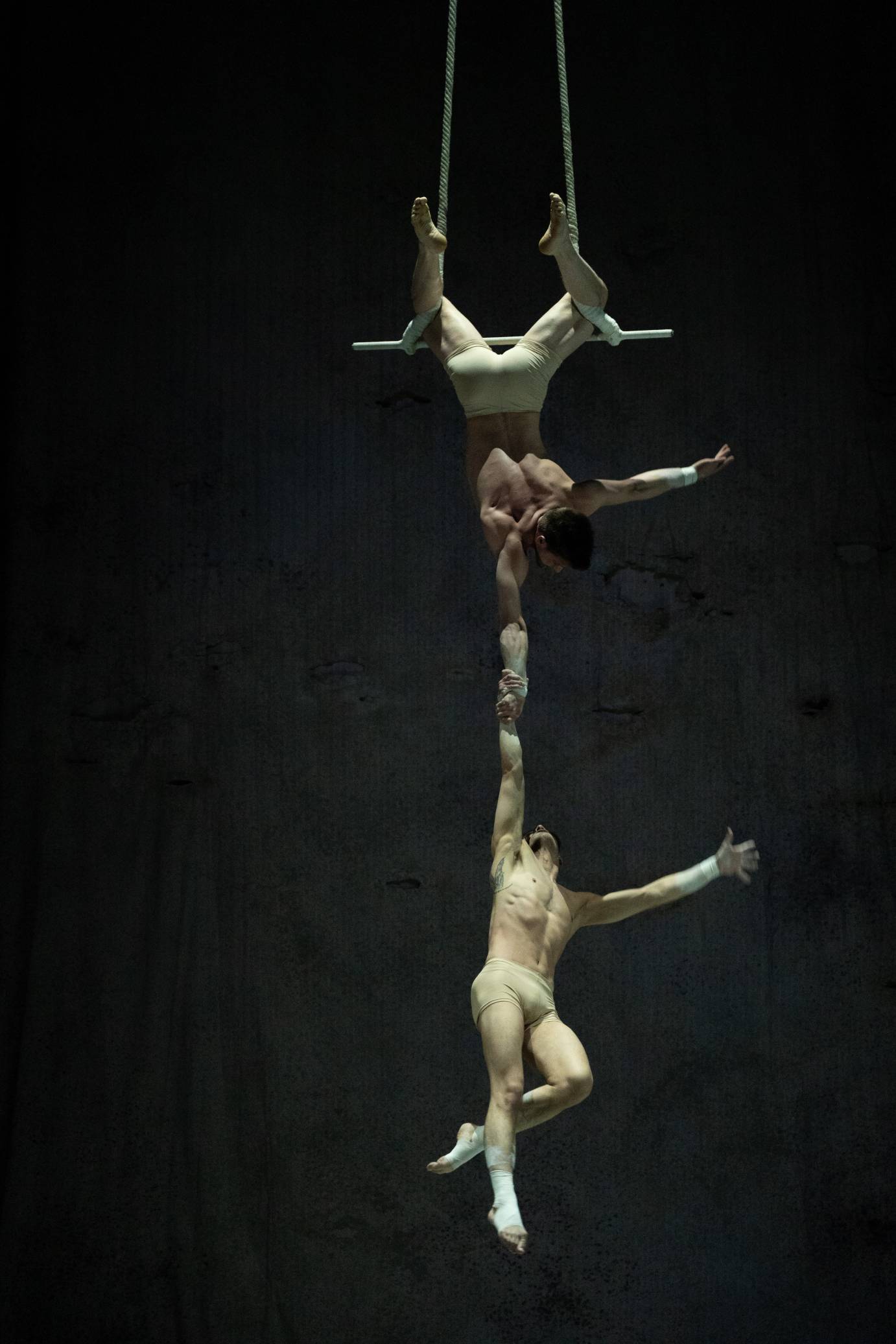 One man hangs from a trapeze holding another man with one hand