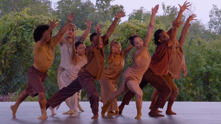 a group of tan and brown dancers against a backdrop of trees, reaching up to the sky