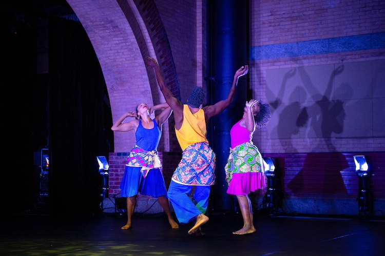 three dancers in bright outfits with gorgeously colorful patterned scarves tied around their waists, form a circle and move together with arms, chests, and faces lifted towards the sky