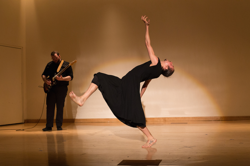 A guitarist plays in the background while a soloist in a black tunic arches his back. His leg hovers in the air and flexes at the ankle.