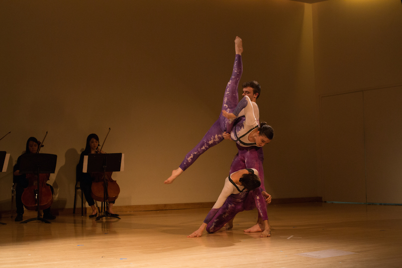 A man lifts a woman in the air as she extends into a penchee arabesque position. Another woman kneels in front of the couple. A group of cellists are playing in the background.
