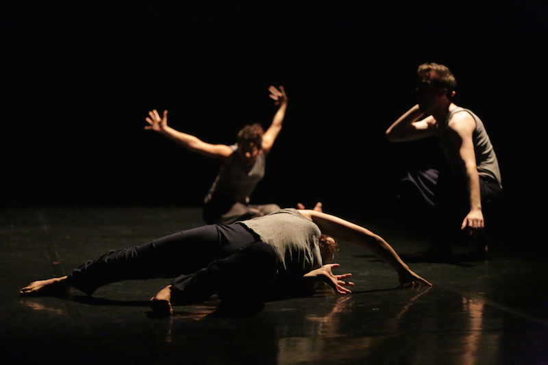 Three dancers in low light in various low lying poses. One dancer in the background splays their arms wide. Another stretches on the floor. The third sits in a crouch.