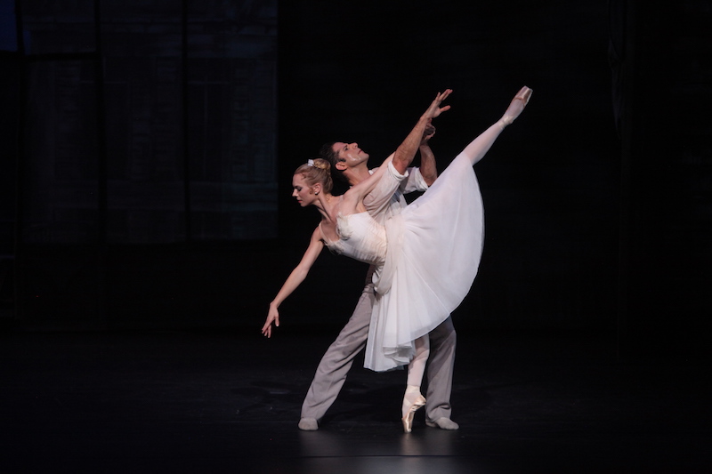 A dancer in white poses in a penchee en pointe while her partner stands behind her.