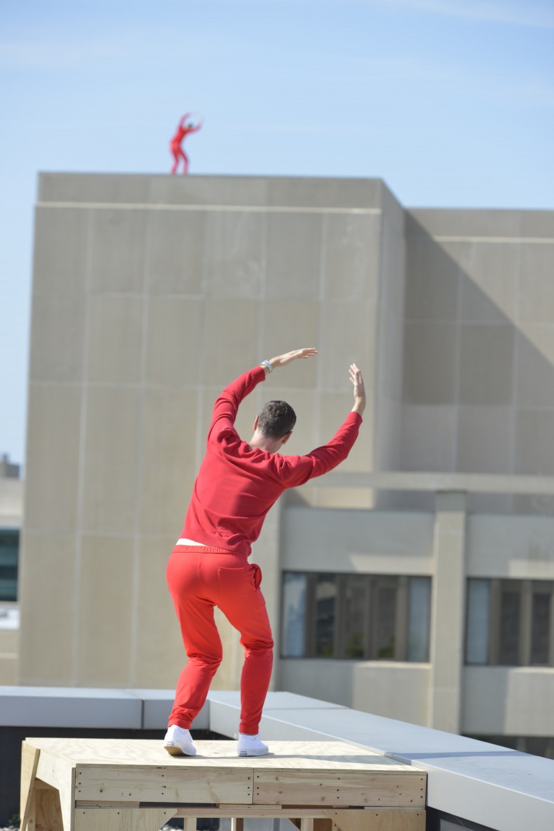 A man on a building in the distance in red. Another man in all red stands on a nearby perch with his back facing the camera.