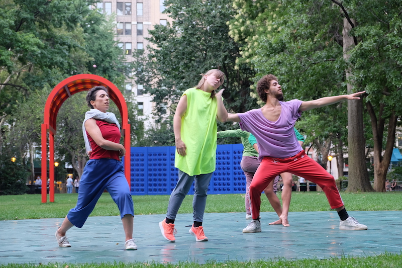 Dancers in primary colors assume varying positions on a prismatic glass and steel floor that sits atop grass