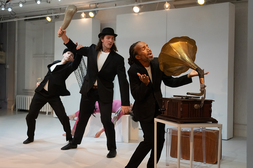 three men, one man in a black suit and pants sings into a victrola, behind him is a man with a club holding it in the air prepared to hit the singer. behind the fellow with a club another similarly dressed man grabs the club swingers elbow . A female body lies on a block behind them... surreal