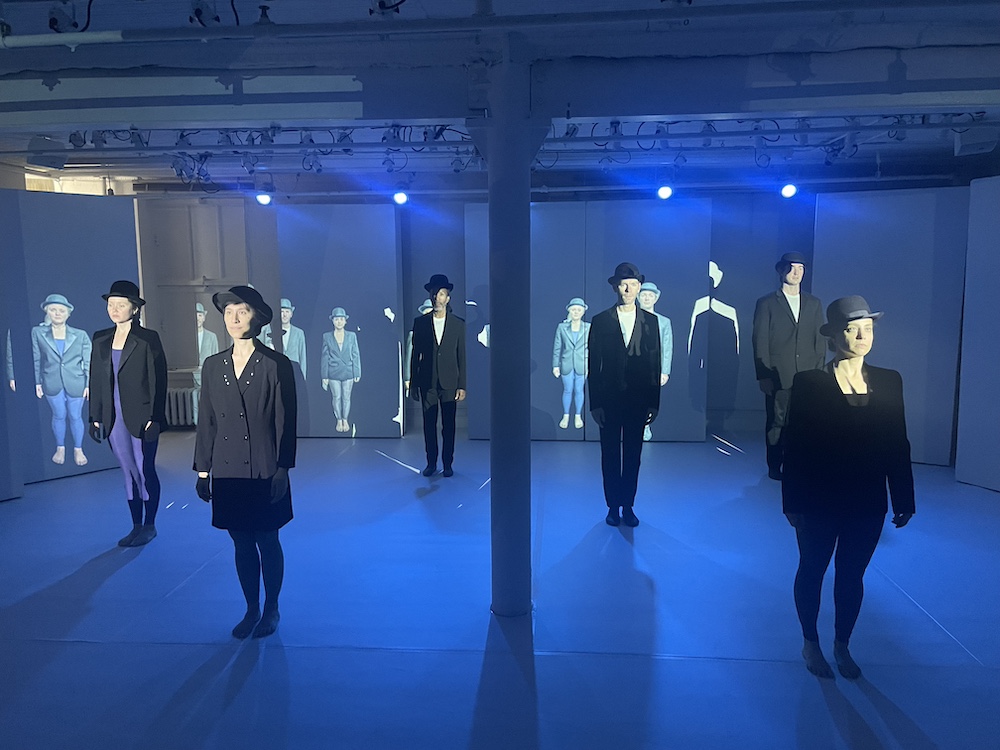 the company stares out at us all wearing black bowlers and black suit jackets behind them are projections of dancers similarly dressed