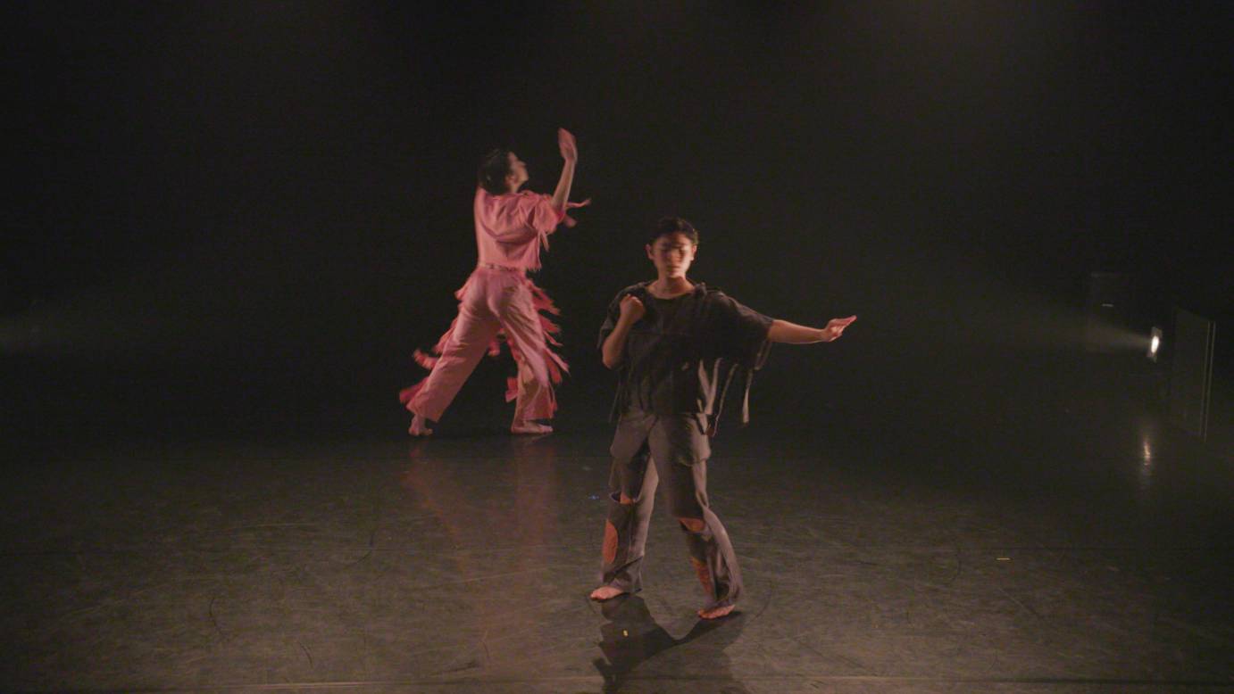 Dancer in all black holds a hand to chest and reaches arm out to side. Dancer in background is dressed in pink and reaches an arm upwards
