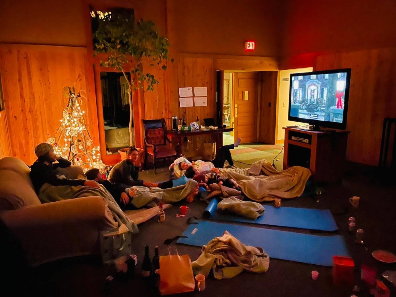 dancers lying on floor watching tv while one sits  in chair a lighted twig christmas tree created by dancers brightens the wood paneled den