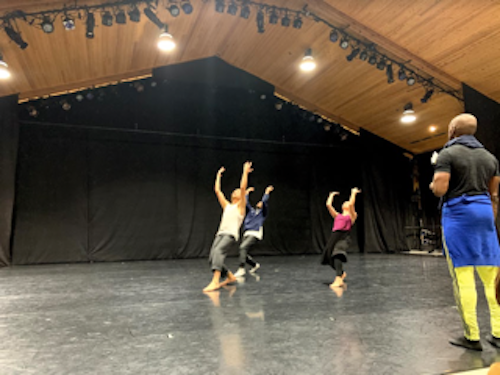 3 dancers in background all wearking dark pants, light tops, no shoes, arms raised in arc towards the high ceiling, bald black dancer in black t shirt with lemon yellow pants and a blue sweatshirt around their waist looking on as dancers work
