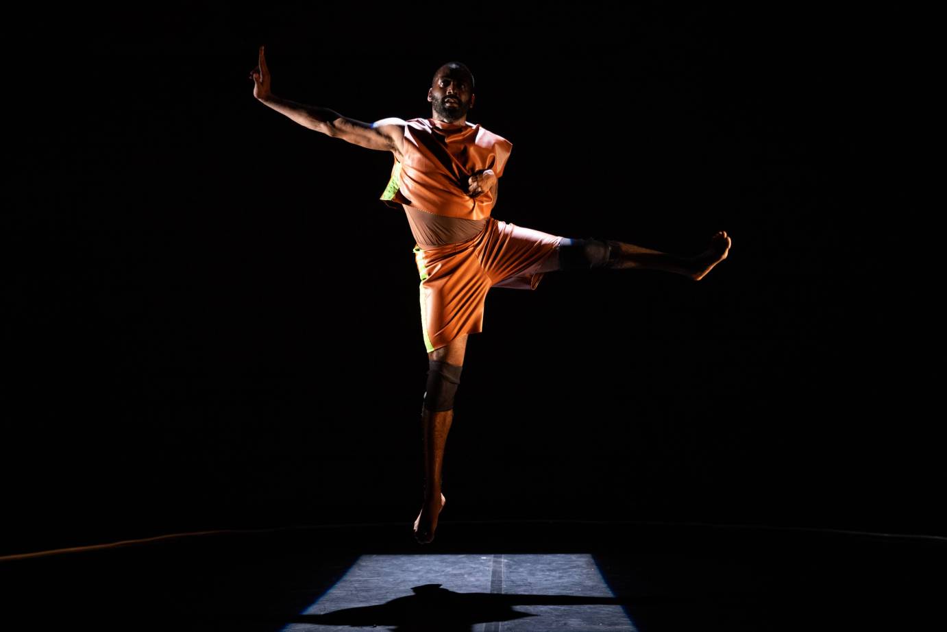 against a black background dancer Jerron Herman, a black man in an orange midriff top and shorts, jumps into the air with one are lifted to his side and the other immobile because of his disability. He is jumping in the air with one leg kicked to the side. 