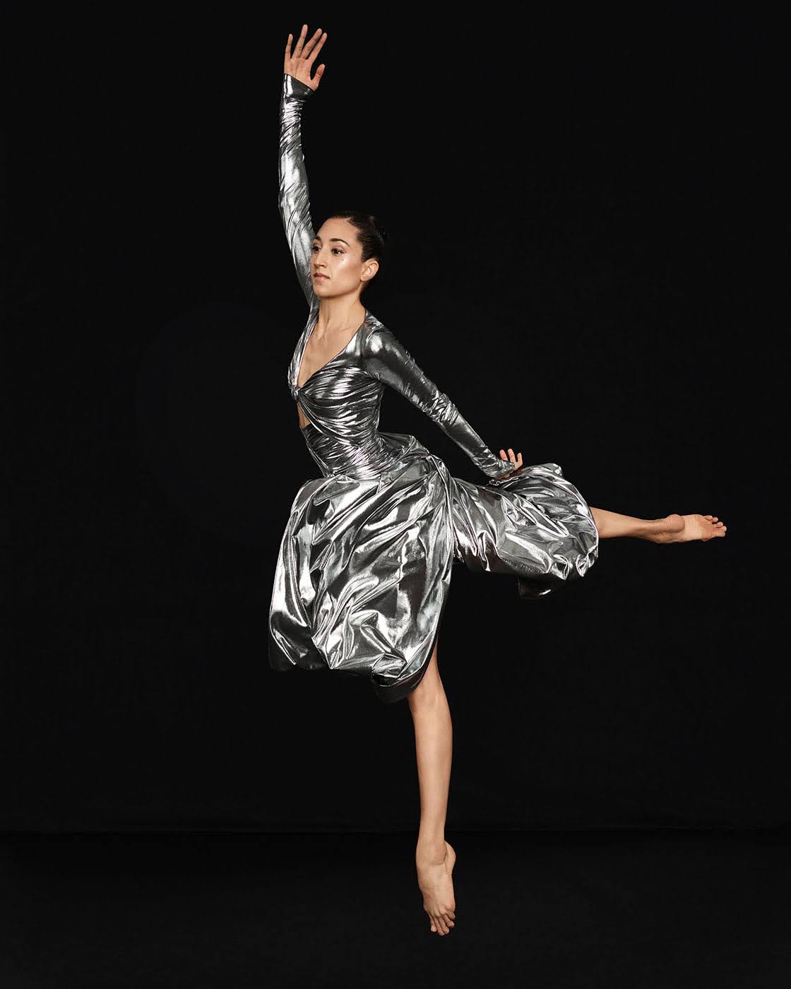 Larissa Asebedo dressed in a gorgeous silver costume with a fitted torso and ballon legs, jumps into the air in a 90 degree arabesque.
