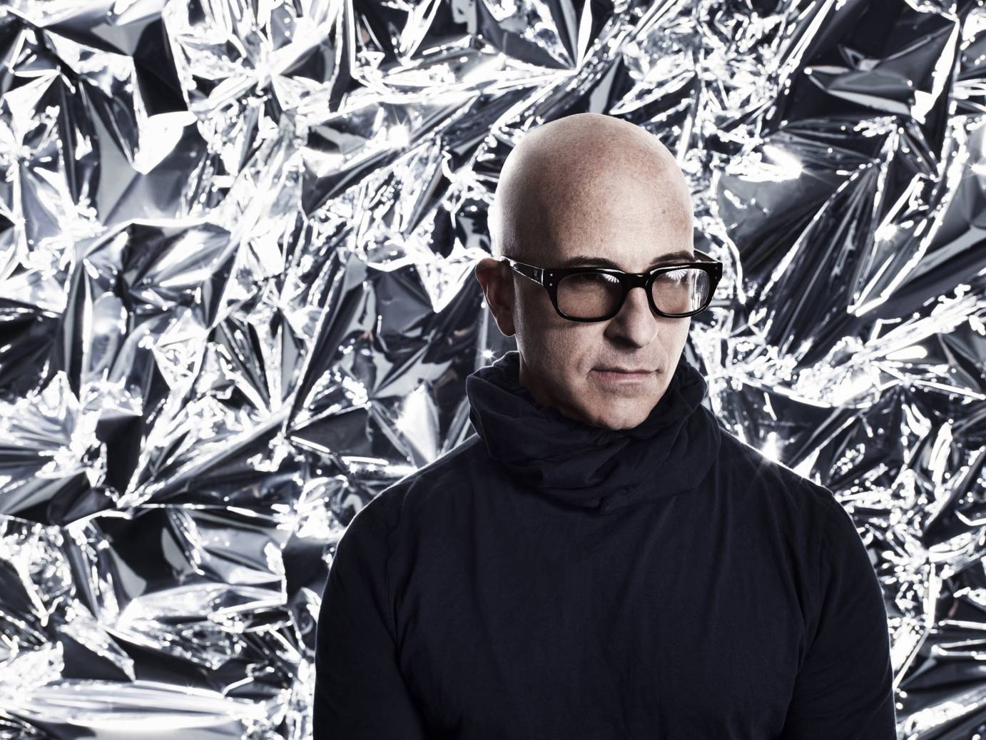 stephen petronio headshot  against a large crinkled aluminium-foil like background. He is bald and wears dark glasses with a dark cableknit turtleneck sweater... he looks down.